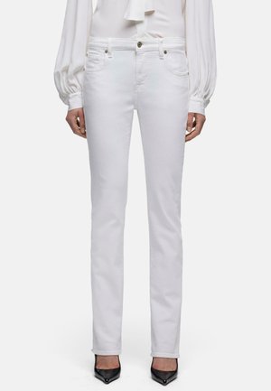Frau, die weiße Slim-Fit-Jeans, eine weiße Bluse mit langen Ärmeln und schwarze spitze High Heels trägt, vor einem schlichten Hintergrund steht.