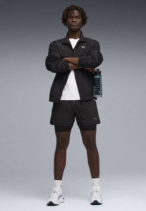 Uomo in piedi con le braccia incrociate, indossa giacca nera Puma e pantaloncini, sneakers bianche, tiene in mano una bottiglia d'acqua blu Puma su sfondo grigio.