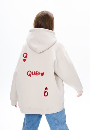 Licht beige oversized hoodie met rode geborduurde tekst "QUEEN" en decoratieve elementen zoals een hart en een ring. Zachte stoftextuur.