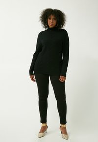 Maglione nero a collo alto lavorato a maglia con motivo a treccia testurizzato, abbinato a leggings neri e tacchi beige lucenti con dettagli dorati.