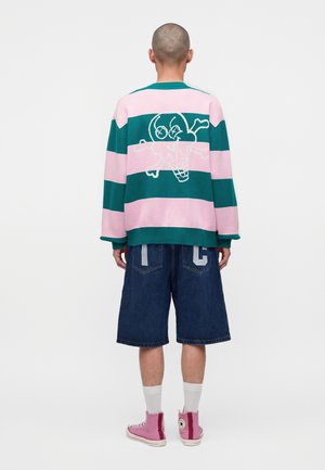 Personne avec la tête rasée portant un pull rayé rose et vert avec un motif de crâne de dessin animé dans le dos, un short en jean, des chaussettes blanches et des baskets roses.