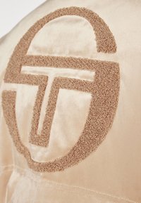 Il tessuto in raso beige presenta un logo marrone testurizzato realizzato con fili tufted. Il design include bordi arrotondati e un motivo di lettere stilizzato e audace.