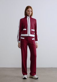 Βουργουνδίας φόρμα με λευκές λεπτομέρειες. Η ζακέτα διαθέτει φερμουάρ και μπροστινές τσέπες, το παντελόνι έχει δύο τσέπες. Ελαφρύ ύφασμα, άνετη γραμμή.