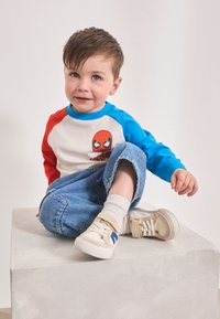 Maglietta bianca a maniche lunghe con maniche blu e rosse e grafica di Spider-Man. Jeans in denim e sneakers beige con cinturini.