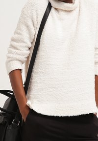 Pull blanc texturé avec col roulé et manches trois-quarts, associé à un sac noir, porté avec un pantalon foncé.