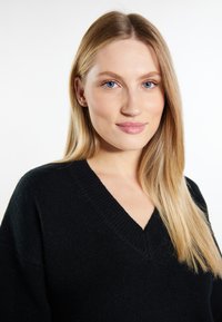 Czarny, dzianinowy sweter z dekoltem w kształcie V i luźnym krojem. Miękka faktura, ribbowe detale przy kołnierzu oraz gładki materiał. Modelka z prostymi, blond włosami.