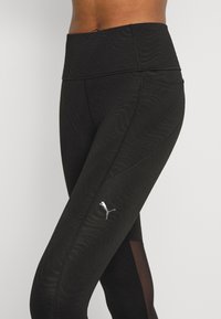 Leggings de entrenamiento negros con un patrón de olas texturizado, cinturilla alta y un pequeño logo plateado en la parte inferior de la pierna con acentos de paneles transparentes.