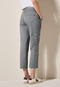 Pantaloni cargo grigi con un fitting slim, lunghezza cropped, multiple tasche tra cui dettagli con zip e una texture liscia. Abbinati a sneaker bianche.