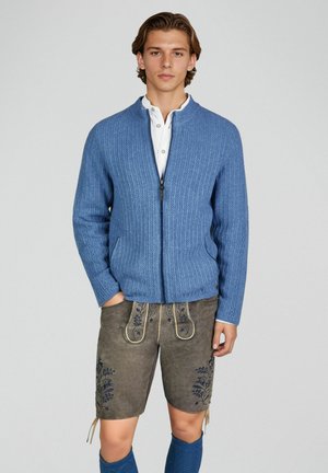 Jeune homme portant un pull en maille bleu zippé, une chemise blanche, un short traditionnel en cuir marron brodé, et des chaussettes assorties bleu jusqu'aux genoux.