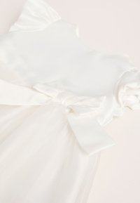 Wit babyjurkje met pofmouwen, satijnen lijfje, tule rok en satijnen strik in de taille, plat gelegd op een lichte ondergrond.