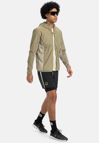 Letthætte i beige med lynlås foran, parret med sorte shorts med grafisk design. Modellen bærer solbriller og sneakers.