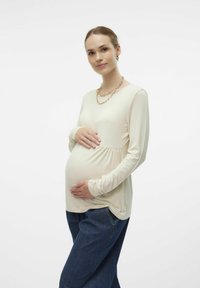 Šviesiai smėlinis nėščiųjų marškinėlis ilgomis rankovėmis, švelniai susiraukšlėjęs po krūtine, derinamas su tamsiai mėlynomis džinsais, vilkimas modelio su matomu nėštumo pilvuku.