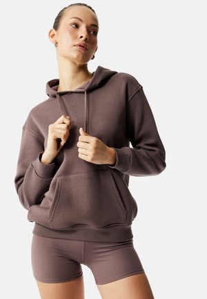 Jonge vrouw met een bruine hoodie en bijpassende korte broek, die de koordjes van de hoodie verstelt, tegen een effen lichte achtergrond.