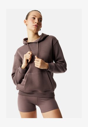 Joven mujer con sudadera con capucha marrón y pantalones cortos a juego, ajustando los cordones de la sudadera, sobre un fondo claro y liso.