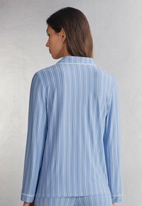 Lichtblauw gestreepte pyjama top met lange mouwen en een klassieke kraag. Heeft witte strepen en subtiele stikdetails op de manchetten en de kraag.