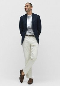 Marineblazer über einem weiß-blau gestreiften T-Shirt, cremfarbene Hose und braune Wildlederschuhe, stehend mit entspannten Armen und lächelnd.