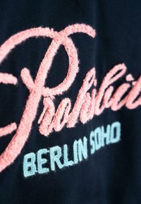 Tamsiai mėlynas audinys, dekoruotas rožiniu išsiuvinėtu užrašymu „Draudžiama“ ir „BERLIN SOHO“ šviesiai mėlyna spalva, su tekstūruotu, kilpinio siūlo finišu.