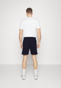 Jūras spēku sporta šorti ar baltām sānu strīpām, elastīga josta, apvienoti ar vienkāršu baltu T-kreklu, modele valkā baltas kedas un zeķes.