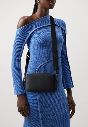 Černá kožená crossbody taška obdélníkového tvaru s uzávěrem na západku a nastavitelným popruhem, nošená přes modré žebrované šaty.