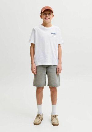 Uśmiechnięty chłopiec w białej koszulce z krótkim rękawem, szarych szortach, beżowych butach z białymi skarpetkami i różowej czapce z daszkiem na jednolitym tle.