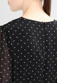Tissu noir avec de petits pois blancs, présentant un col rond et une fermeture éclair discrète au dos. Les manches transparentes ajoutent de la texture.