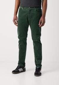 Man som bär mörkgröna manchesterbyxor och svarta Adidas-sneakers med vita ränder, stående mot en vit bakgrund.