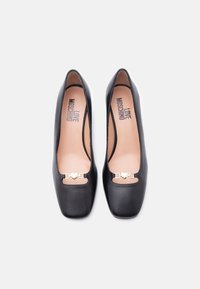 Ζευγάρι μαύρες δερμάτινες μπαλαρίνες με τετράγωνη μύτη, λεπτομέρεια με αλυσίδα σε σχήμα χρυσής καρδιάς και λογότυπο "LOVE MOSCHINO" στο εσωτερικό, προβολή από πάνω σε λευκό φόντο.