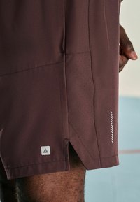 Bordeaux atletikshorts med perforerede sidepaneler, med et hvidt logo patch og subtile zigzag-dekorationer i kanten. Glat stofstruktur.