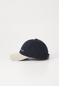 Marineblå kasket med en beige skygge. Har et tekstureret stof og justerbar rem. Brandnavn vist med hvidt på forsiden.