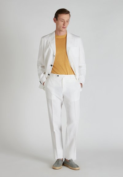 Blazer bianco in lino con bottoni neri, pantaloni coordinati e una maglietta in maglia giallo senape. Scarpe grisette slip-on completano l'outfit.