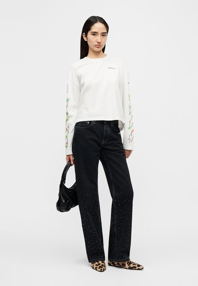 Άσπρο μακρυμάνικο πουκάμισο με πολύχρωμα ανθισμένα σχέδια στα μανίκια, συνδυασμένο με μαύρα τζιν με στρας στο τελείωμα και παπούτσια με ζώα μοτίβο.