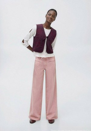 L'outfit a strati presenta un gilet trapuntato borgogna con chiusura a legatura sopra una camicia bianca a maniche lunghe e pantaloni larghi rosa. Le scarpe marroni completano il look.