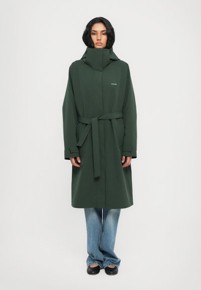 Dark green long coat with a high collar, a belt, and loose-fitting sleeves. Paired with blue flared jeans and black slip-on shoes.