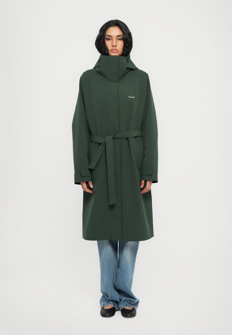 Manteau long vert en matériau lisse, avec un col haut, une ceinture et des poches latérales, associé à un jean bleu évasé et des chaussures noires.