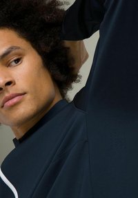 Top blu navy a maniche lunghe con colletto alto, caratterizzato da una texture liscia e dettagli bianchi a contrasto. Piccole perforazioni sono visibili sulla manica.