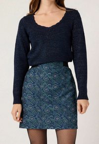 Marinblå stickad tröja med en scalloped halsringning, i kombination med en blommönstrad grön och blå kjol. Strukturerad finish och nätstrumpbyxor.