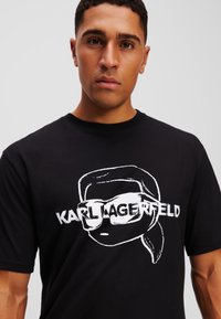 Melna kokvilnas T-kreklā ar baltu grafiku, kurā attēlots karikatūras tēls ar saulesbrillēm un zem tā teksts "KARL LAGERFELD". Apaļais dekoltē.