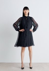 Schwarzes Kleid mit Punkten, durchsichtigen langen Ärmeln, Knopfleiste und Bindeband am Hals. A-Linien-Form, knielang. Glatte Stofftextur.