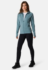 Light blue zip-up jacket with textured fabric, high collar, and two side pockets, paired with black fitted pants and black athletic shoes.