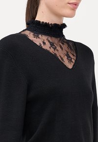 Maglione nero con inserti in pizzo trasparente sulla scollatura, con colletto arricciato. Texture a costine e design a forma di V ne valorizzano l'aspetto.