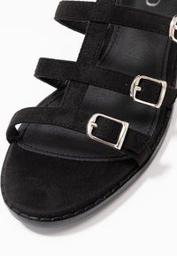 Musta mokkasaapas, jossa on useita ristiin meneviä hihnoja ja hopeisia solkia, sileä musta pohja ja teksturoitu pohja.