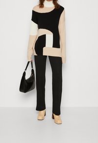 Strickpullover mit abstrakten Mustern in Schwarz, Weiß und Beige; schwarze Ripp-Hosen; beige Blockabsatzschuhe; schwarze Tasche mit geschwungener Form.