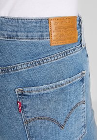 Persona que lleva unos vaqueros Levi's azules con el bolsillo trasero visible, parche de cuero de la marca y etiqueta roja de Levi's en la costura del bolsillo.