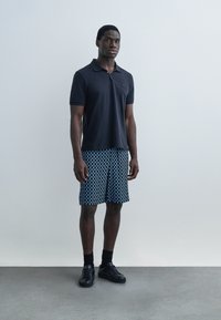 Mladi moški stoji v črni polo majici, modrih kratkih hlačah z vzorcem, črnih nogavicah in črnih supergah na enostavni svetli podlagi.