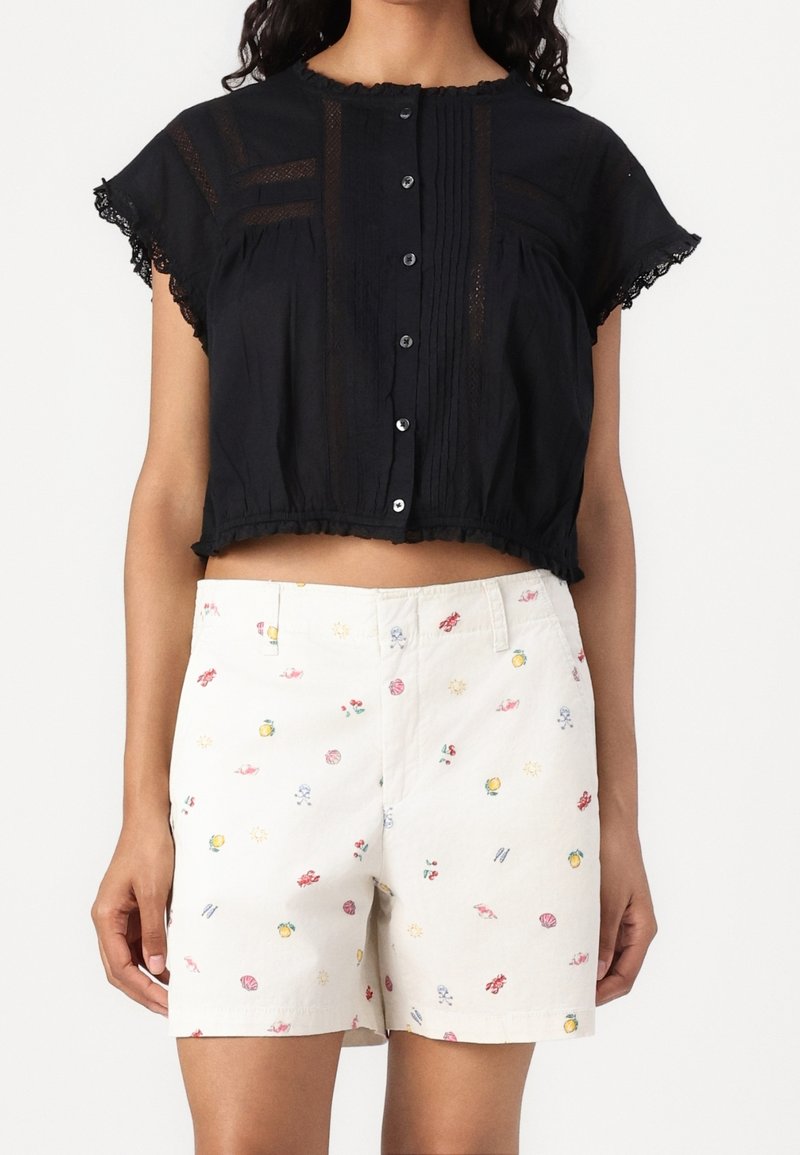 Top corto nero con chiusura a bottone e bordi in pizzo; abbinato a pantaloni corti beige con una stampa colorata di frutta e motivi.