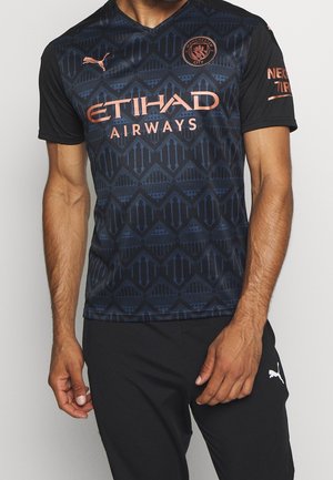 Mand iført sort og marineblå Manchester City fodboldtrøje med kobberlogoer og sorte Puma bukser mod en ensfarvet baggrund.