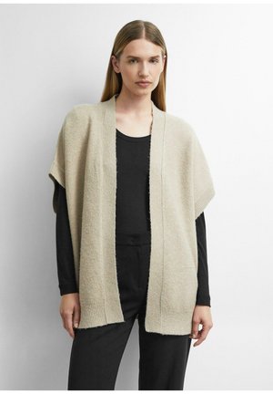 Woman with straight hair wears a beige open-front knit vest over a black long-sleeve shirt and black pants, standing against a white background.