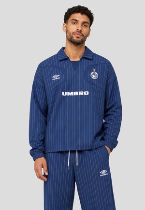 Mann, der ein blau gestreiftes Umbro Langarmshirt und passende Hose trägt, steht mit einer Hand in der Tasche vor einem grauen Hintergrund.