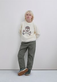 Bluză crem cu imprimeu de craniu și oase încrucișate, asortată cu pantaloni verde măsliniu și adidași albastru-gri cu benzi Velcro.