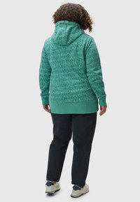Haut à capuche turquoise avec un motif texturé, capuche à cordon de serrage et ourlet vert uni. Porté avec un pantalon noir et des baskets de couleur claire.